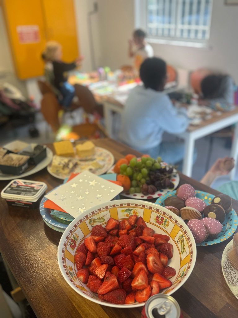 A table of party food, strawberries, iced cup cakes, grapes and cheese. In the background children are doing craft activities at another table.