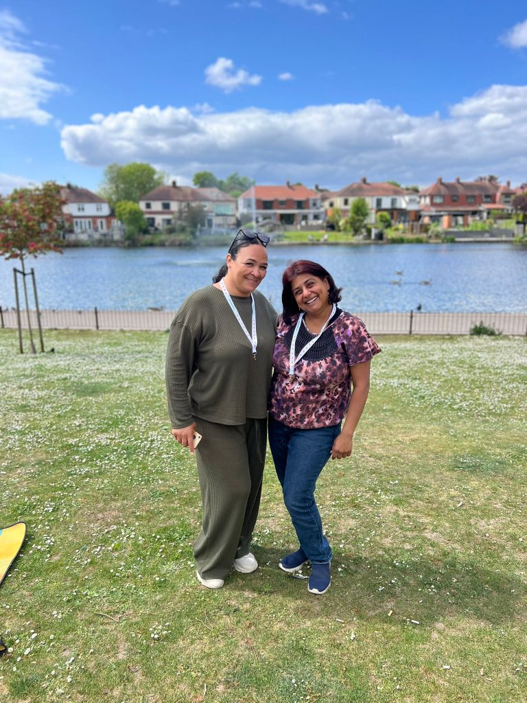 Hema and Sarah are standing side by side smiling at the camera, on a sunny day.