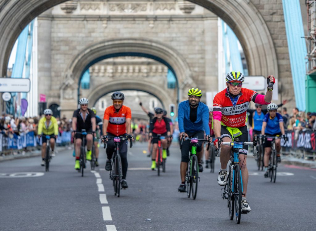 Cyclists ride across a road bridge, past a watching crowd