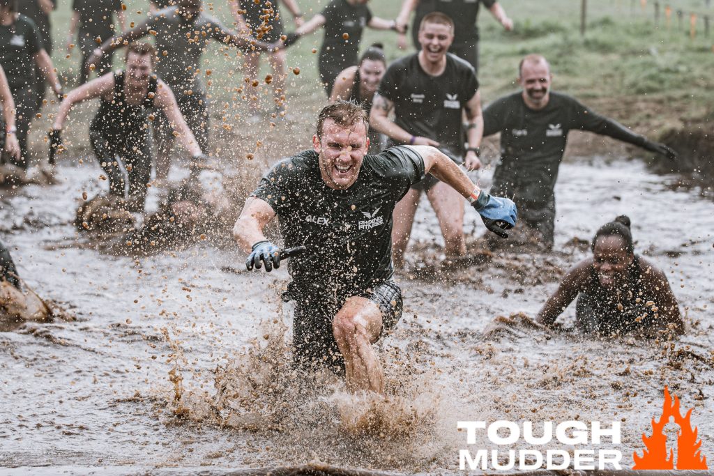 Competitors at a Tough Mudder event, running through muddy water. Their faces are exhilarated and determined.